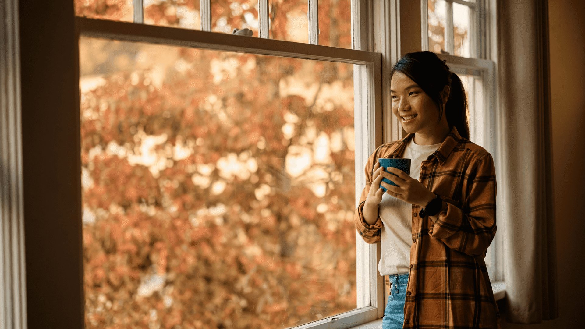 A woman smiles while holding a mug and looking out her window, with fall foliage in the background, symbolizing seasonal home preparation and fall pest control.